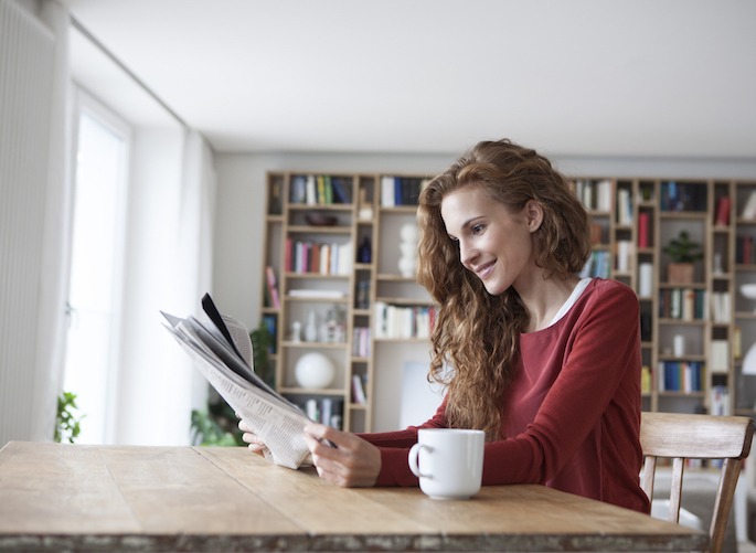 newsroom-header Smiling woman at home sitting at wooden table with cup reading newspaper